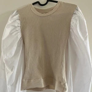 Beige ribbad topp med puffärmar - Trendig topp med ribbad beige kropp och kontrasterande vita puffärmar. Rund halsringning och figurnära passform. Perfekt för dig som vill sticka ut med en cool och stilren look.