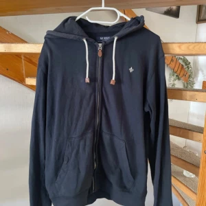 Svart hoodie med dragkedja från Morris - Svart hoodie från Morris med vit snörning i huvan och dragkedja framtill. Klassisk passform med två fickor och liten broderad logga på bröstet. Perfekt för chill dagar eller när du vill ha en enkel och stilren look.