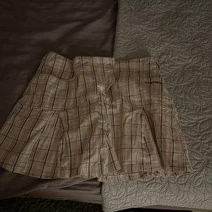 Kjol med inbyggd shorts!  - Beige och ljusrosa rutig kortkjol från Cross med veckade detaljer och fickor framtill. Kjolen har bälteshällor och broderad logga på ena fickan. Perfekt för en sportig och trendig look. Kjolen har även shorts inbyggt. OBS strl 40 men liten i storleken!🩷