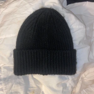 Svart ribbstickad mössa från Lindex - Svart ribbstickad mössa från Lindex med uppvikt kant. Tillverkad i en mjuk blandning av återvunnen polyester som håller dig varm och snygg under kyliga dagar. Enkel och stilren design som passar till allt.