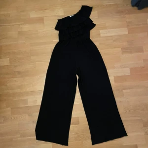 Svart byxdress med volang från Kappahl - Svart byxdress från Kappahl i storlek 152. Modellen har en axel, volangdetaljer upptill och vida ben. Materialet är mjukt och lätt, perfekt för en trendig look. Sitter snyggt i midjan med resår och har en stilren, enkel design.