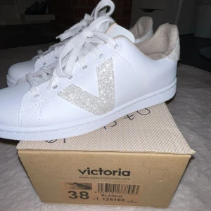 Vita sneakers Victoria med glitterdetaljer - Snygga vita sneakers från Victoria med glittrande silverdetaljer på sidan och hälen. Skorna har klassisk rund tå, snörning och platt sula. Dem är helt nya och aldrig använda, säljer pågrund att dem var för små för mig och även är lite små i storlek, skulle säga att skon passar till någon med storlek 37-37,5 och inte 38🤍🤍ursprungs pris - 687!!!