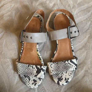 Sandaler med ormskinnsmönster och klack - Snygga sandaler från Bianco med breda remmar i beige skinn och coolt ormskinnsmönster i svart och vitt. Skorna har en låg klack med samma mönster och justerbart spänne vid ankeln. Perfekta för att lyfta din sommarstil.