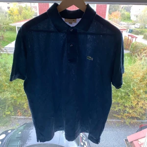 Marinblå pikétröja från Lacoste - Snygg marinblå pikétröja från Lacoste med klassisk krage, knappar och den ikoniska krokodilloggan på bröstet. Tröjan är i 100% bomull och har korta ärmar. Perfekt för dig som gillar stilren och sportig look.
