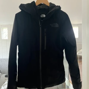 Svart skidjacka från The North Face - Superfin svart skidjacka från The North Face med huva och bröstficka med dragkedja och snölås.