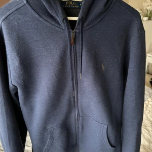 Marinblå hoodie från Polo Ralph Lauren - Snygg marinblå hoodie från Polo Ralph Lauren med dragkedja framtill, huva med snörning och klassisk logga broderad på bröstet. Tröjan har känguruficka och är tillverkad i mjuk bomullsmix, perfekt för chill dagar.