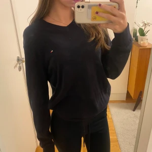 Mörkblå v-ringad tröja Tommy Hilfiger - Mörkblå v-ringad tröja från Tommy Hilfiger med liten broderad logga på bröstet. Tröjan är i mjuk bomull och har långa ärmar samt ribbade muddar vid ärmslut och nederkant. Unisex