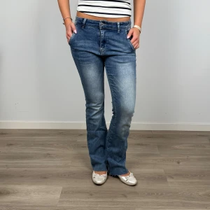 Blå bootcut jeans med låg midja - Snygga blå jeans med bootcut passform och låg midja. Jeansen har klassisk femficksdesign, bälteshällor och subtila slitningar framtill och baktill. Materialet är mjukt denim som ger en bekväm känsla och en avslappnad look.