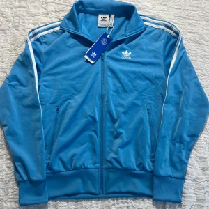Blå Adidas track jacket med vita ränder - Snygg blå Adidas track jacket med klassiska vita ränder längs ärmarna och broderad logga på bröstet. Jackan har dragkedja framtill, hög krage och två fickor med dragkedja. Tillverkad i polyester och perfekt för en sportig streetstyle.