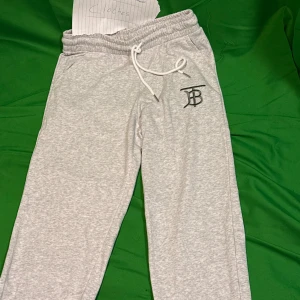 Grå mjukisbyxor med snörning - Snygga ljusgrå mjukisbyxor med elastisk midja och vit snörning. Byxorna har en svart broderad logga på ena benet och är tillverkade i mjukt bomullsmaterial. Perfekta för en chill och avslappnad stil.Liknande modell som originalet!