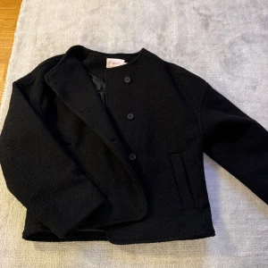 Svart ulljacka från Nelly - Stilren svart jacka från Nelly i ullblandning med rak passform och knäppning framtill. Jackan har diskreta fickor på sidorna och en enkel, kraglös design som ger en clean look. Perfekt för dig som gillar minimalistisk stil.