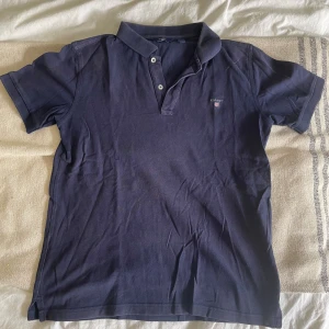 Marinblå pikétröja från Gant - Säljer en klassisk marinblå pikétröja från Gant med broderad logga på bröstet. Tröjan har korta ärmar, krage och två knappar framtill. Tillverkad i mjuk bomull som är skön att bära hela dagen.