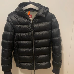 Svart pufferjacka Parajumpers Sheen M - Svart pufferjacka från Parajumpers, modell Sheen, med huva och dragkedja framtill. Jackan har ett stort broderat PJS-märke på ärmen och ribbade muddar vid ärmslut och nederkant. Perfekt för kalla dagar och riktigt snygg streetstil. Finns ett hål fram till men annars i väldigt fint skick kan gå ner i pris vid snabb affär!!!