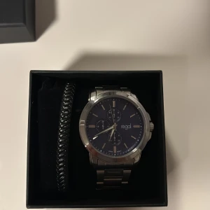 Regal klocka med blå urtavla och länk - Snygg klocka från Regal med marinblå urtavla och silverfärgad länk i metall. Klockan har flera små urtavlor och stilrena indexmarkeringar. Levereras i originalask med extra länkbitar och svart armband i flätad design.