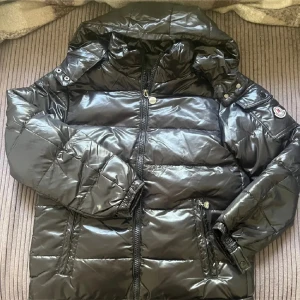 Svart pufferjacka med huva från M Fashion  - Snygg svart pufferjacka från M Fashion med glansig finish, huva och dragkedja framtill. Jackan har M Fashion -logga på ärmen, praktiska fickor med dragkedja och quiltad design. Perfekt för kalla dagar när du vill hålla stilen.