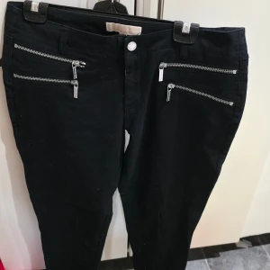 Svarta byxor Michael Kors med dragkedjor - Svarta byxor från Michael Kors med fyra snygga silverfärgade dragkedjor framtill. Byxorna har en slim passform och är tillverkade i ett stretchigt bomullsmaterial. Perfekta för dig som gillar detaljer och vill sticka ut lite extra. Nypris kring 2000 kr
