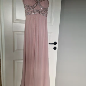 Ljusrosa långklänning med strass - Ljusrosa långklänning från Y&C med tunna axelband och vackra strassdetaljer i korsmönster över bysten. Klänningen har ett luftigt, svepande tyg och en elegant, figurnära överdel. Perfekt för dig som vill sticka ut med glittriga detaljer och romantisk känsla.