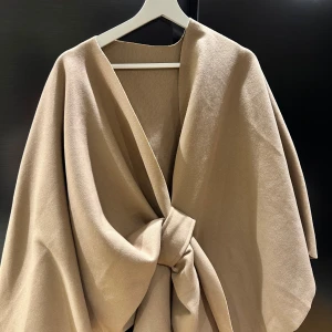 Beige omlott kappa med knyt - Snygg beige kappa i omlottmodell med breda ärmar och knyt framtill. Jackan har en stilren och minimalistisk look, perfekt för dig som gillar en clean vibe. Materialet är mjukt och känns lyxigt mot huden. Passar dig som vill ha en trendig och enkel jacka till höst och vår.