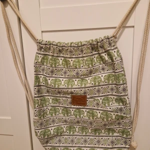 Thai väska thai tum - Snygg tygryggsäck med grönt elefantmönster och etniska detaljer på vit botten. Dragskostängning med grova rep och en brun läderpatch framtill. Perfekt för dig som gillar unika accessoarer med boho-vibe.