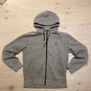 Grå hoodie från Polo Ralph Lauren - Säljer en grå hoodie från Polo Ralph Lauren med dragkedja och svart logga på bröstet. Tröjan har huva med snörning, två fickor framtill och är tillverkad i mjuk bomullsmix. Perfekt för chill dagar eller när du vill ha en clean streetstyle.