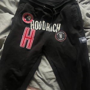 Svarta Hoodrich mjukisbyxor med patchar - Svarta mjukisbyxor från Hoodrich med stora broderade patchar och tryck framtill. Byxorna har snörning i midjan, ribbade muddar vid benslut och flera detaljer i rött, vitt och svart. 