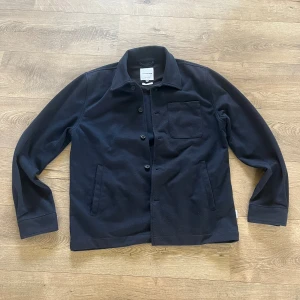 Svart overshirt från Lindbergh - Stilren svart overshirt från Lindbergh i regular fit. Jackan har klassisk krage, knäppning framtill, en bröstficka och två sidofickor. Materialet är mjukt och flexibelt, perfekt för lager-på-lager. Enkel och clean look som funkar till det mesta.