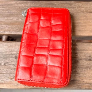 Röd plånbok i skinn från Wera - Snygg röd plånbok från Wera i präglat skinn med croco-look. Plånboken har dragkedja runtom, flera kortfack och ett myntfack med dragkedja på baksidan. Perfekt för dig som vill ha en stilren och trendig accessoar med plats för allt du behöver. Oanvänd, fint skick! Längd: 15cm Bredd:10cm