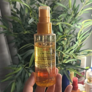 Nuxe Sun Moisturizing Protective Oil - Fuktgivande och skyddande olja för håret från Nuxe Sun. Kommer i en genomskinlig plastflaska med pump och guldorange detaljer. Innehåller 100 ml och skyddar mot UV-strålar, salt och klor. Perfekt för sol-exponerat hår. Använd 
