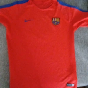 FC Barcelona Nike fotbollströja röd - Säljer en röd FC Barcelona fotbollströja från Nike med blå detaljer på axlarna och klubbmärke broderat på bröstet. Tröjan har korta ärmar, BEKO-tryck på ryggen och är tillverkad i lätt och ventilerande material. Perfekt för dig som älskar Barca och vill sticka ut på planen.