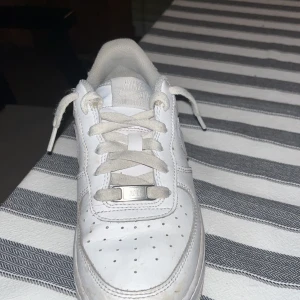 Nike Air Force 1 vita sneakers - Klassiska Nike Air Force 1 sneakers i helvitt skinn med vit sula och snörning. Ikonisk design med perforerad tå och Nike swoosh på sidorna. Perfekta för dig som gillar en clean och tidlös stil.