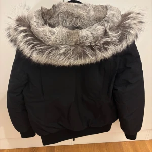 Svart dunjacka med päls från Mackage - Säljer en svart dunjacka från Mackage med stor fluffig fuskpäls runt huvan. Jackan har dragkedja, ribbade muddar och flera fickor med dragkedja. Perfekt för kalla vinterdagar och riktigt snygg streetstil.