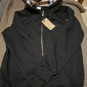 Svart hoodie från Burberry med dragkedja - Svart hoodie från Burberry med klassiskt rutigt foder i huvan. Tröjan har dragkedja framtill, snörning i huvan och två fickor. Broderad Burberry-logga på bröstet. Perfekt för dig som vill ha en stilren och lyxig streetwear-look. En stor storlek M.