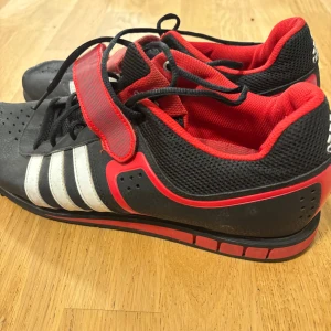 Svarta och röda Adidas sneakers - Säljer ett par svarta och röda Adidas sneakers med vita ränder på sidan. Skorna har snörning och kardborreband, samt mesh- och syntetmaterial för extra andningsförmåga. Adidas-logga på hälen och röd detalj längs sulan. Perfekta för dig som gillar sportig stil.