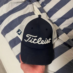 Titleist keps mesh Pro V1 blå/vit - Snygg keps från Titleist med mörkblå front och skärm samt vit mesh baktill. Broderad Titleist-logga framtill och Pro V1-text på sidan. Justerbar med kardborreband. Aldrig använd men inga lappar eller kvitto kvar (inga defekter) 