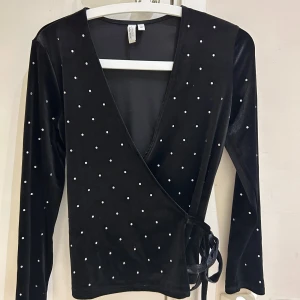 Ny Svart omlottopp i sammet från & Other Stories - Supersnygg svart omlottopp i sammet från & Other Stories med långa ärmar och små glittriga pärldetaljer över hela toppen. Djup v-ringning och knytband i sidan ger en trendig look. Perfekt för dig som vill sticka ut lite extra.