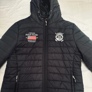 Svart pufferjacka från Vinson Polo Club - Svart pufferjacka från Vinson Polo Club med broderade loggor på bröstet och amerikansk flagga. Jackan har huva med snörning, dragkedja framtill och två fickor med dragkedja. Perfekt för kyliga dagar och riktigt snygg streetstil. Knappt använd..max 2 gång
