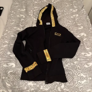 Svart/Guld EA7 kofta - Svart koftafrån EA7 Emporio Armani med guld logga på bröstet och guldiga band med tryck på huva och ärmslut. Dragkedja framtill och ficka med dragkedja. Skön passform. Går ej att köpa från butik. Lite slitage i ärmarna.