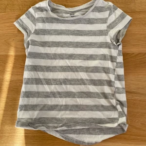 Randig t-shirt från H&M Basic - Kortärmad t-shirt från H&M Basic i ekologisk bomull. T-shirten har breda horisontella ränder i vitt och ljusgrått, rund halsringning och en klassisk passform. Perfekt för en avslappnad stil.