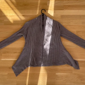 Grå ribbad kofta med lång ärm - Säljer en grå ribbad kofta med lång ärm och öppen front. Koftan har en snygg, avslappnad passform och är perfekt att slänga på sig när det är lite kyligare. Materialet är mjukt och stretchigt, vilket gör den superbekväm.