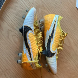 Nike Mercurial Vapor 360 fotbollsskor gul elit  - Säljer ett par Nike Mercurial Vapor 360 elite fotbollsskor i storlek 40. Skorna är i en snygg gul och vit färg med svarta detaljer och har snörning. Ovandelen är i syntetmaterial och designen är riktigt sportig med en stor Nike-logga på sidan. Perfekta för dig som vill sticka ut på planen.