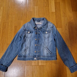 Blå jeansjacka från Detroit by Lindex - Snygg blå jeansjacka från Detroit by Lindex med klassisk design, knappar framtill och två bröstfickor med lock. Jackan har långa ärmar och relativt kort passform.