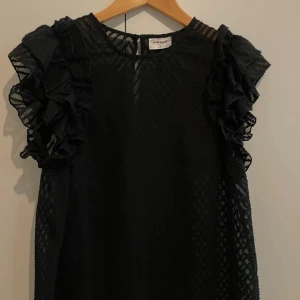 Svart volangklänning från Aware - Supersnygg svart kortklänning från Aware by Vero Moda med transparenta partier och coolt mönster. Klänningen har volangdetaljer på axlarna och är ärmlös. Perfekt för dig som gillar statement-plagg med extra edge.