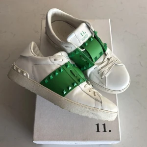 Valentino Rockstud - Snygga Valentino Garavani Rockstud sneakers i vitt skinn med grönt band och ikoniska nitar. Klassisk rund tå, snörning och platt sula. Grön logga på plösen och coola detaljer längs sidorna. Perfekta för dig som vill sticka ut med exklusiva sneakers.
