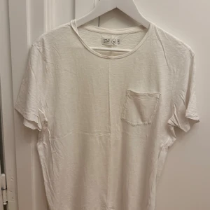 Vit t-shirt från Emilio med ficka - Vit t-shirt från Emilio i mjuk bomull. Klassisk modell med rund halsringning och en liten bröstficka på vänster sida. Perfekt basic-plagg med avslappnad passform och korta ärmar. Enkel och stilren design som funkar till allt. Storlek M.