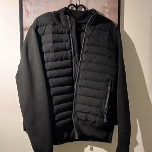 Svart quiltad jacka från Moncler - Säljer en svart quiltad jacka från Moncler med ribbade muddar och dragkedja framtill. Jackan har Moncler-logga på ärmen, två fickor med dragkedja och en stilren, sportig look. Perfekt för dig som vill ha en snygg och bekväm jacka med exklusiv känsla.