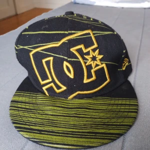 DC svart keps med gul logga - Snygg DC keps i svart med gul broderad logga framtill och bak. Kepsen har gröna och gula ränder över hela och platt skärm med coolt mönster. Perfekt för dig som gillar streetwear och vill sticka ut med en unik stil.