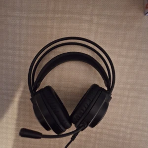 HP Omen Mindframe Gaming Headset - HP Omen Mindframe gaming headset med kabelanslutning, justerbar mikrofon och bekväma over-ear-kuddar. Headsetet är i gott skick utan synligt slitage. Levereras med 3,5 mm och USB-kablar för flexibel anslutning till PC och konsol.