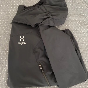 Svart Regn jacka från Haglöfs - Svarta täckbyxor från Haglöfs med dragkedjor vid fickorna och elastisk midja. Perfekta för vinterns alla äventyr och outdoor-aktiviteter. Snygg minimalistisk design med Haglöfs-logga på benet. Materialet är slitstarkt och vattenavvisande syntet. Funkar ochså för tjejer!!