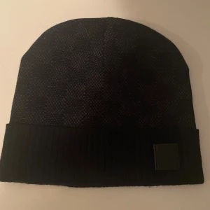 Svart mössa från Louis Vuitton - Snygg svart mössa från Louis Vuitton med diskret rutmönster och ribbad uppvikt kant. På kanten sitter en svart patch med Louis Vuitton Paris-logga. Perfekt accessoar för att lyfta din vinterstil.