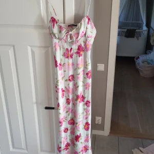 Blommig långklänning från Gina Tricot - Supersöt långklänning med tunna axelband från Gina Tricot. Klänningen är vit med stora rosa och röda blommor, har snörning vid bysten och en mjuk, flowy passform. Perfekt för sommarens alla tillfällen och ger en fräsch vibe. Använd 1 gång.